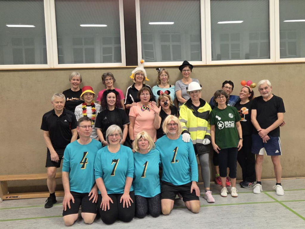 Gruppe von Sportlern in bunten Kostümen, versammelt in einer Sporthalle. Einige tragen sportliche Kleidung, andere haben Kostüme mit Hüten und Accessoires.